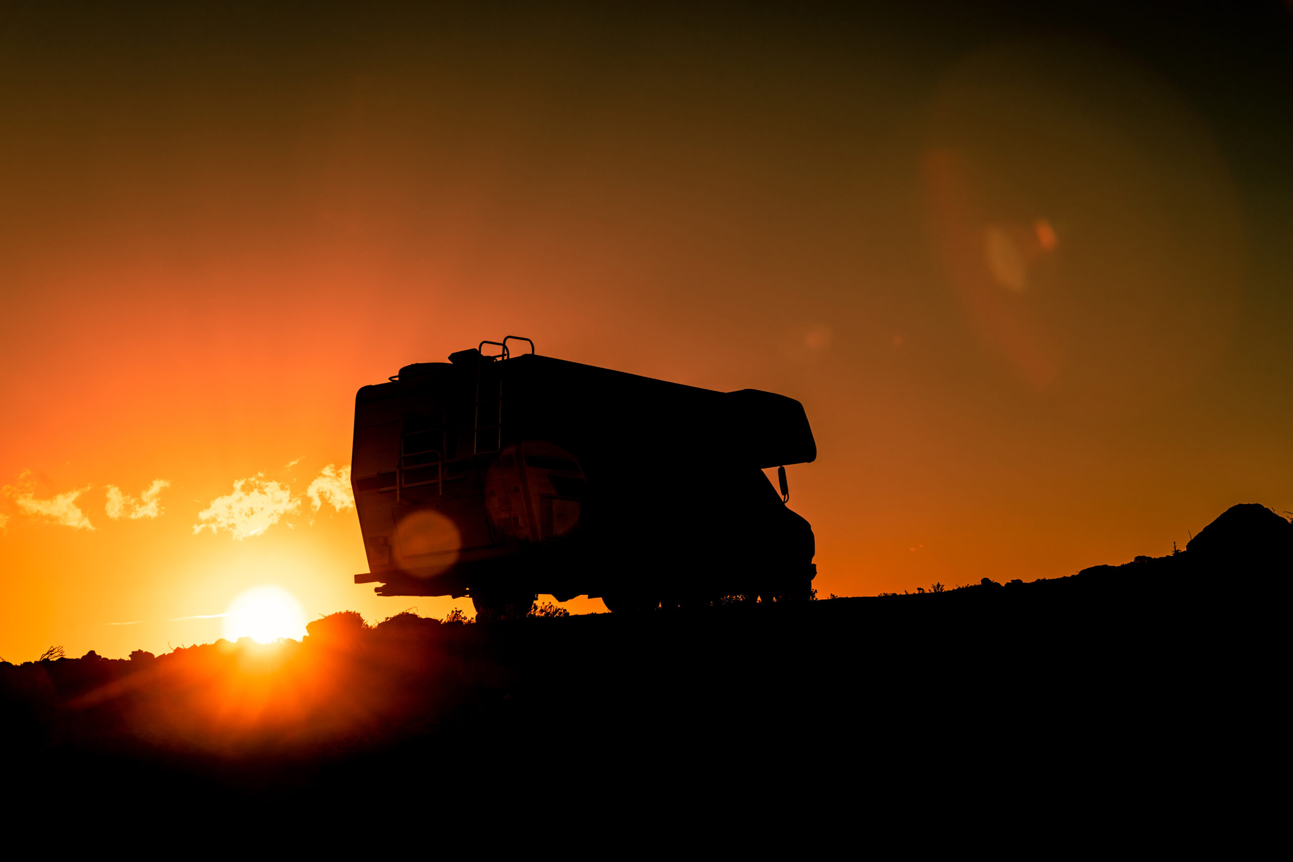A silhouette of a large truck is seen on a hill at sunset, with an orange sky and the sun low on the horizon, casting a warm glow and long shadows.